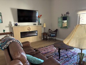 A living room with a brown leather sofa, a flat-screen TV mounted above a fireplace, a patterned rug, and a small table with a lamp. - Winter in AZ!! Early bird pricing!! 1000.00 DOLLARS OFF for any 2 month stay!! (Scottsdale)