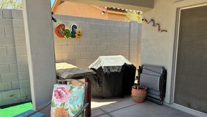 Een terras buiten, met een overdekte barbecue, een stoel met bloemenmotief bekleding, en een bordje waarop 'Cafe' staat.
