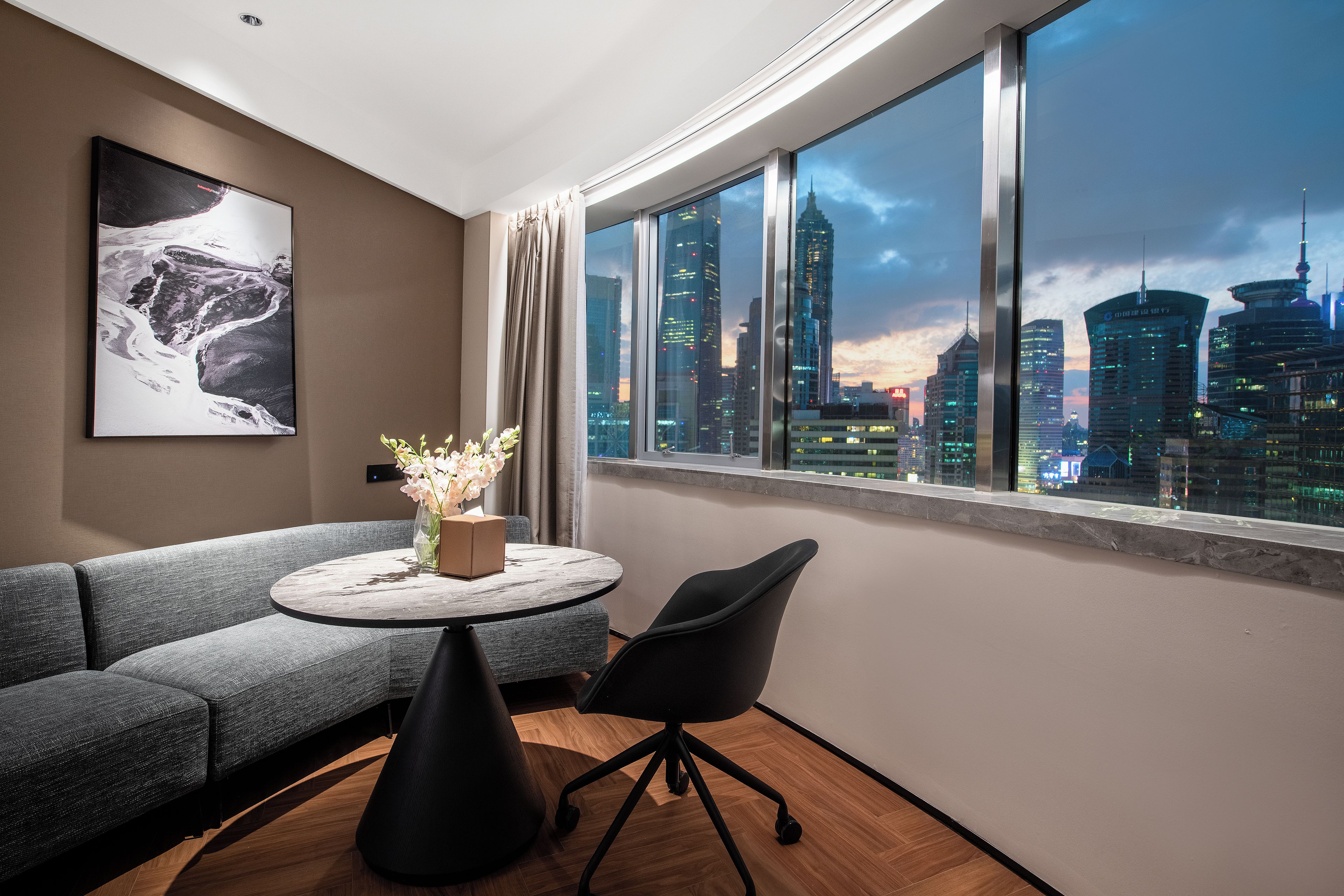 A modern room with a large window, a sofa, a round table with a vase, and a painting on the wall.