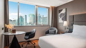 A hotel room with a bed, a desk, a chair, a lamp, and a view of a cityscape through large windows.