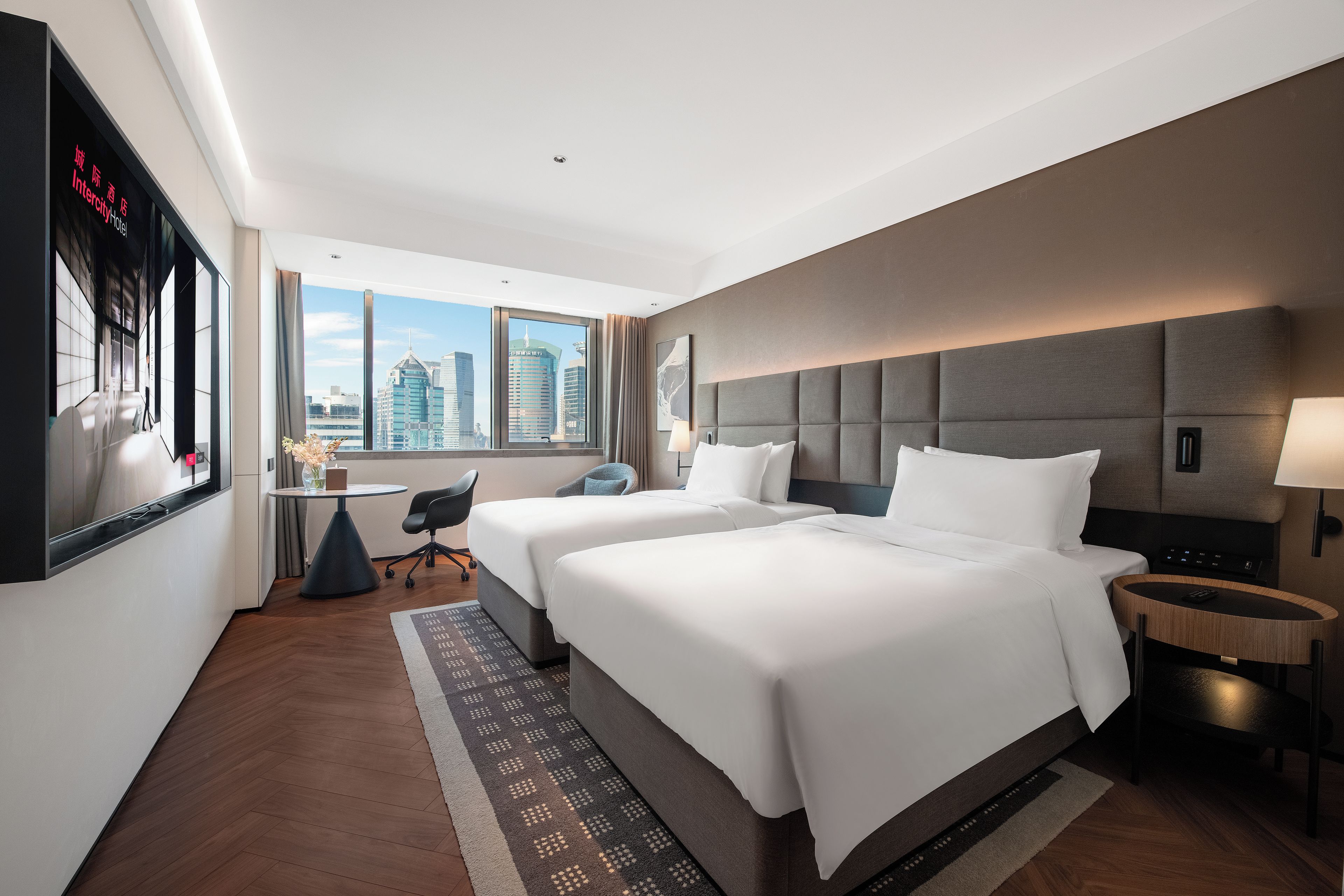 Quarto de hotel com duas camas, uma escrivaninha, uma cadeira, uma televisão e vista para a paisagem urbana.