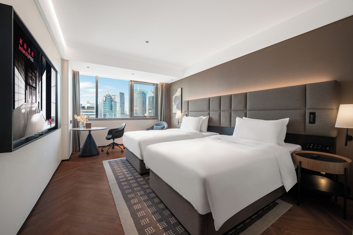 Ein Hotelzimmer mit zwei Betten, einem Schreibtisch, einem Sessel und Blick auf die Stadt.