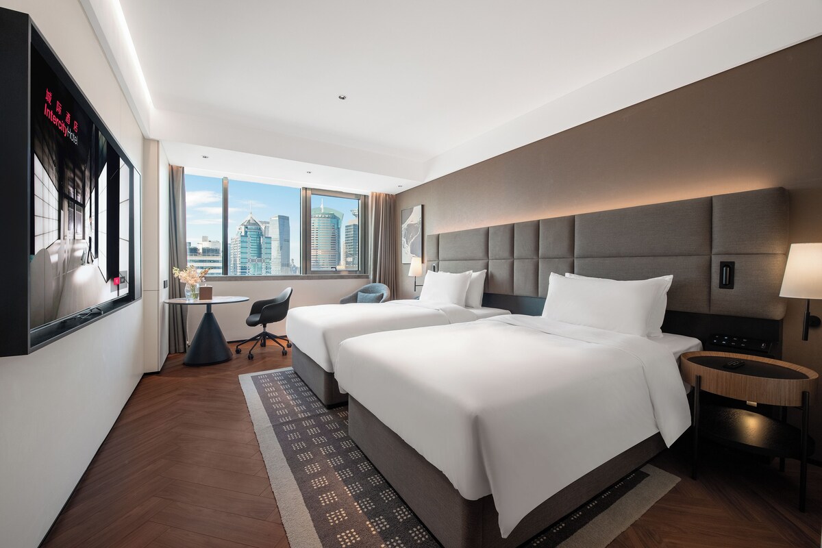 A hotel room with two beds, a desk, a chair, a TV, and a view of the cityscape.
