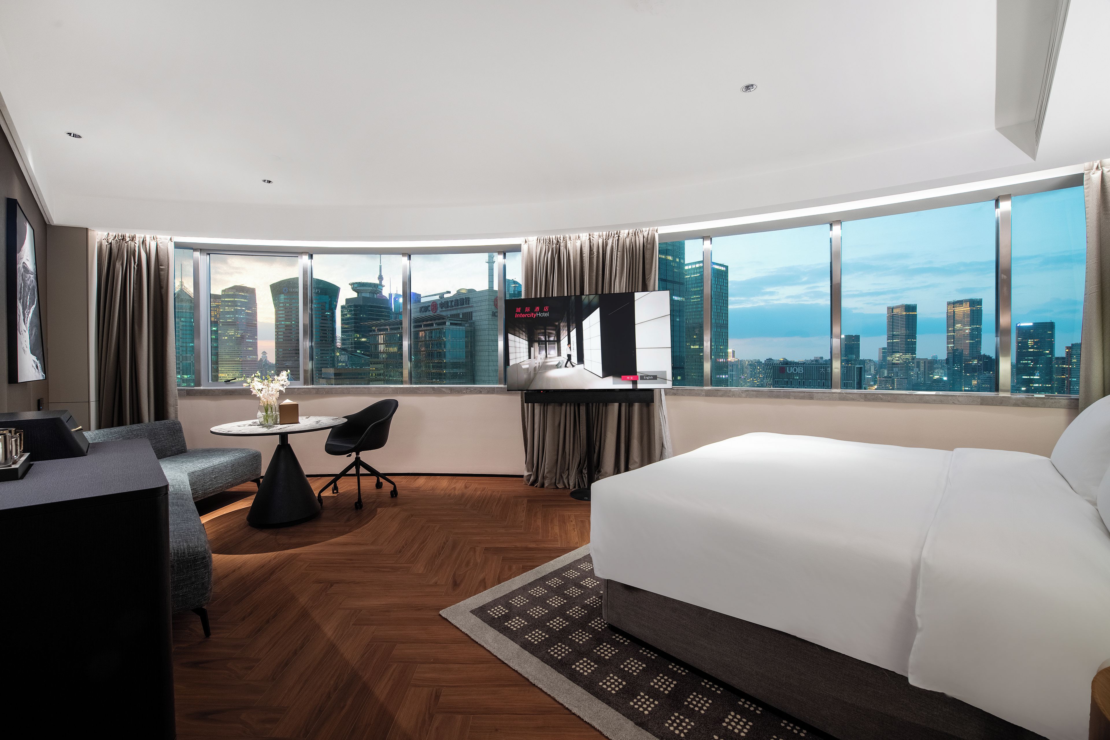 A hotel room with a large bed, a desk with a chair, a small table with a vase of flowers, and a view of the cityscape through large windows.
