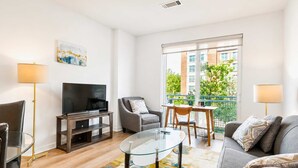 A modern living room with a flat-screen TV, a glass coffee table, a grey sofa, and a balcony with a view of buildings and greenery. - Luxury Apt Downtown with Pool & Gym (Stamford, CT)