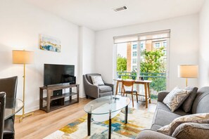 Ein modernes Wohnzimmer mit Fernseher, Glastisch, grauem Sofa und einem Balkon mit Blick auf Gebäude und Grünflächen.