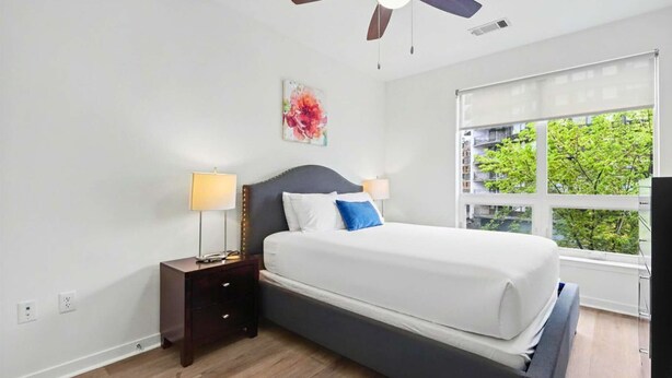 A bedroom with a bed, two bedside tables, a ceiling fan, a window with a view, and a wall-mounted artwork. - Luxury Apt Downtown with Pool & Gym (Stamford, CT)