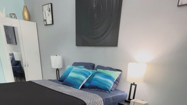 A bedroom with a bed, bedside tables, a desk, a chair, a lamp, a wall art piece, and a wardrobe. - Butterfly Cove (Lead Vale)