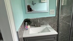A bathroom with a white sink, a mirror, a shower, and a dark gray cabinet. - Butterfly Cove (Lead Vale)