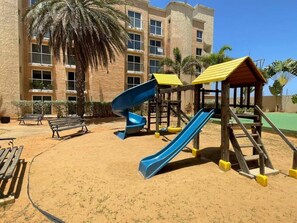 Ein Spielplatz mit Rutschen und Bänken, eine Palme und ein Gebäude im Hintergrund.