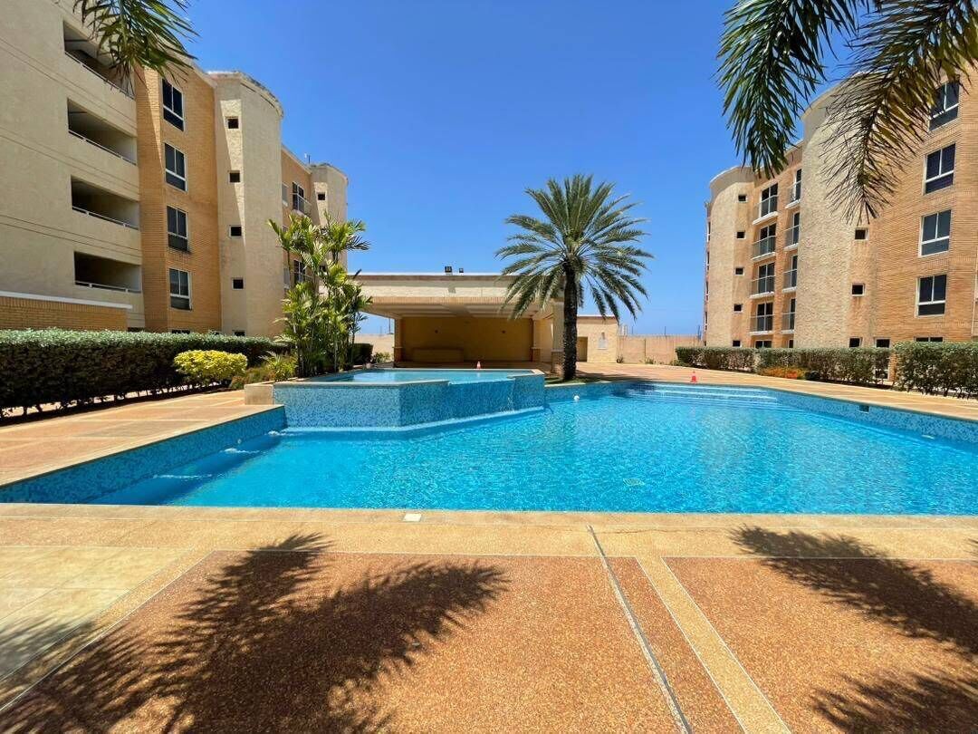 Una piscina con superficie d'acqua blu trasparente, circondata da una zona pavimentata con palme e un edificio sullo sfondo con numerosi balconi.