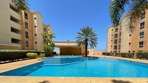Une piscine à l’eau d’un bleu limpide, entourée d’une zone pavée agrémentée de palmiers et, au fond, d’un bâtiment doté de nombreux balcons.