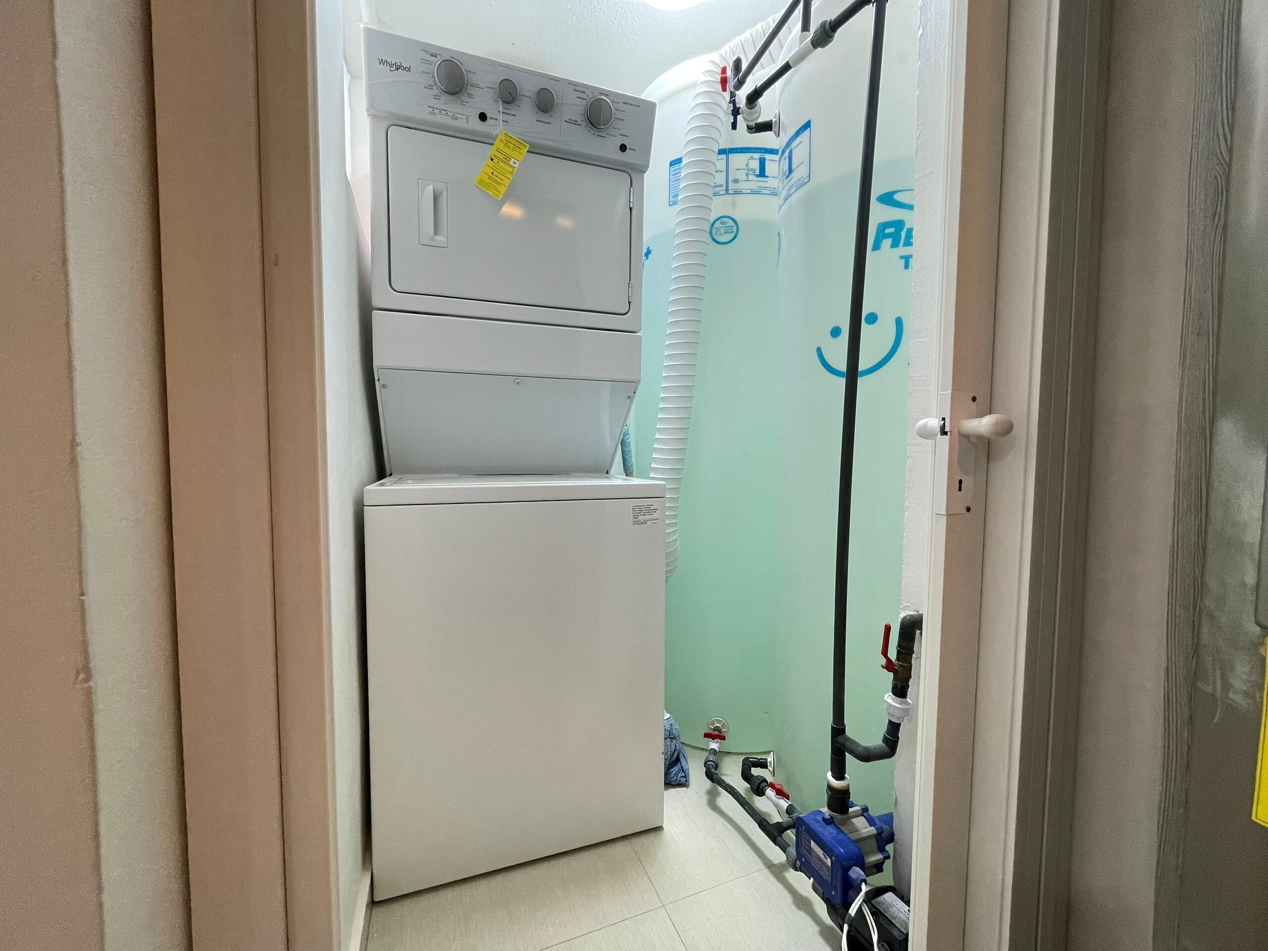 Uma lavanderia com máquina de lavar e secadora Whirlpool, aquecedor de água e encanamento aparente.