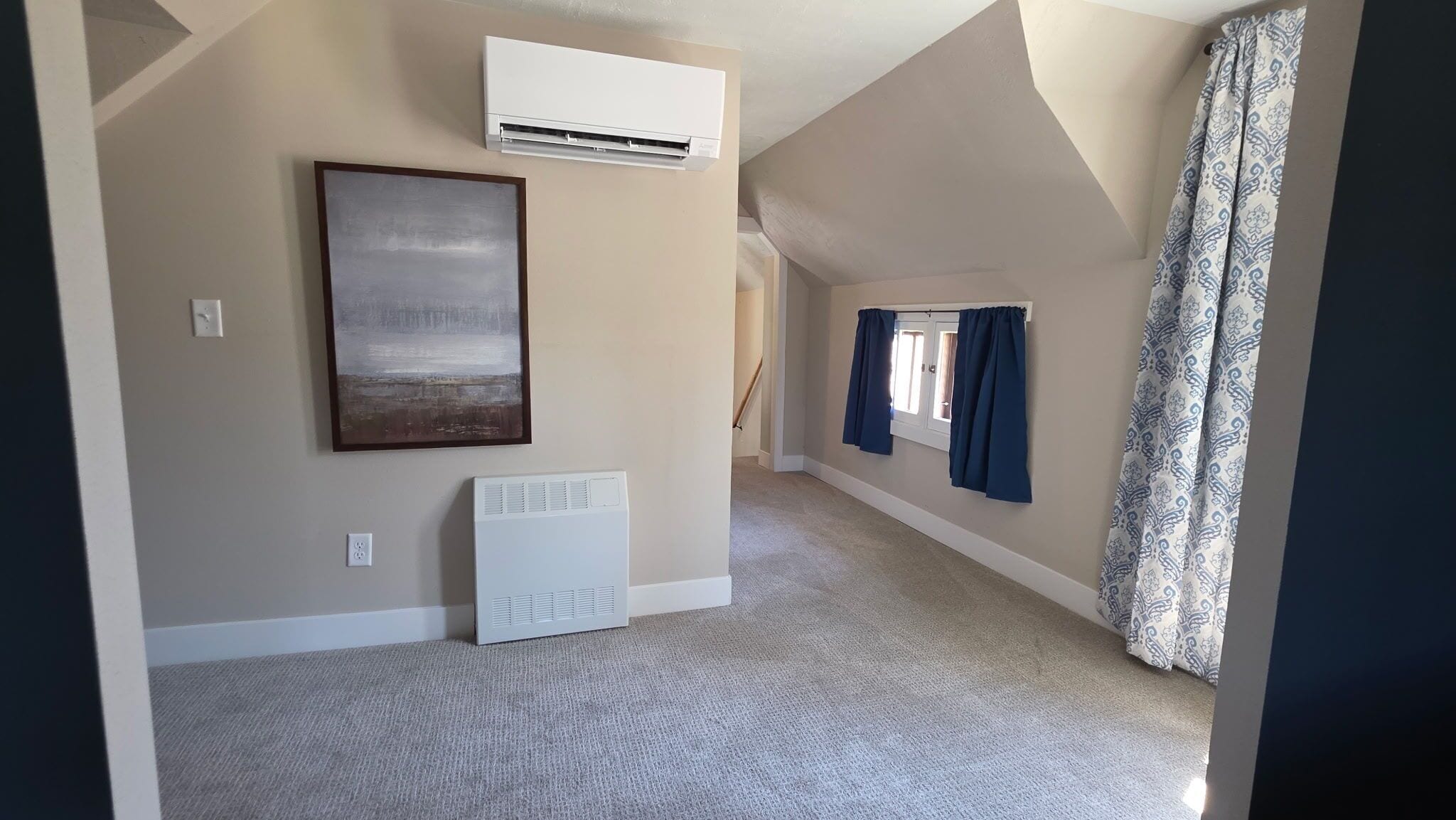 A room with an air conditioning unit, a radiator, a framed picture, and a window with curtains.