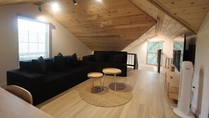 Een moderne loft-achtige woonruimte met een hoge plafond, houten balken, een zwarte modulaire bank, twee kleine ronde tafels en een groot raam dat natuurlijk licht binnenlaat.