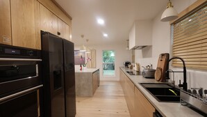 Een moderne keuken met houten kastjes, een zwarte koelkast, een ingebouwde oven, een centraal kookeiland met een spoelbak en hangende lampen.