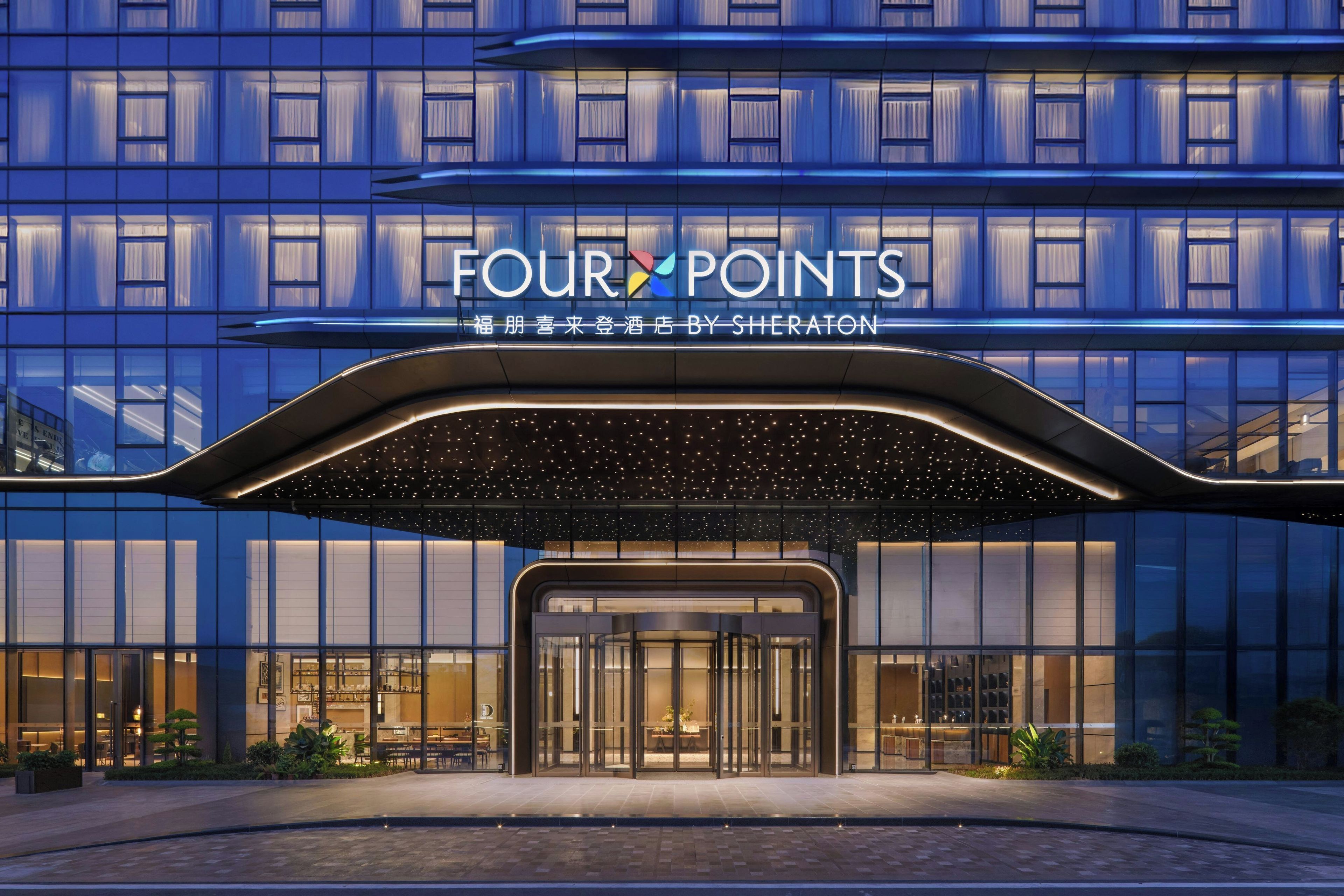 A hotel entrance with a modern design, featuring a glass facade and a sign that reads 'FOUR POINTS BY SHERATON'.