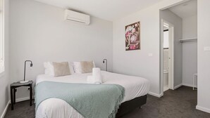 A bedroom with a bed, bedside tables, an air conditioning unit, a painting, and a door leading to a bathroom. - Domi Rentals - The Ormond House (Clayton)