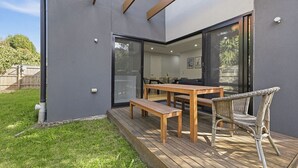 A modern patio with a wooden table and chairs, a glass door leading to an interior space, and a grassy yard. - Domi Rentals - The Ormond House (Clayton)
