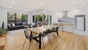 A modern kitchen with white cabinets, a dining area with a wooden table and chairs, and a living area with a sofa and a TV. - Domi Rentals - The Ormond House (Clayton)