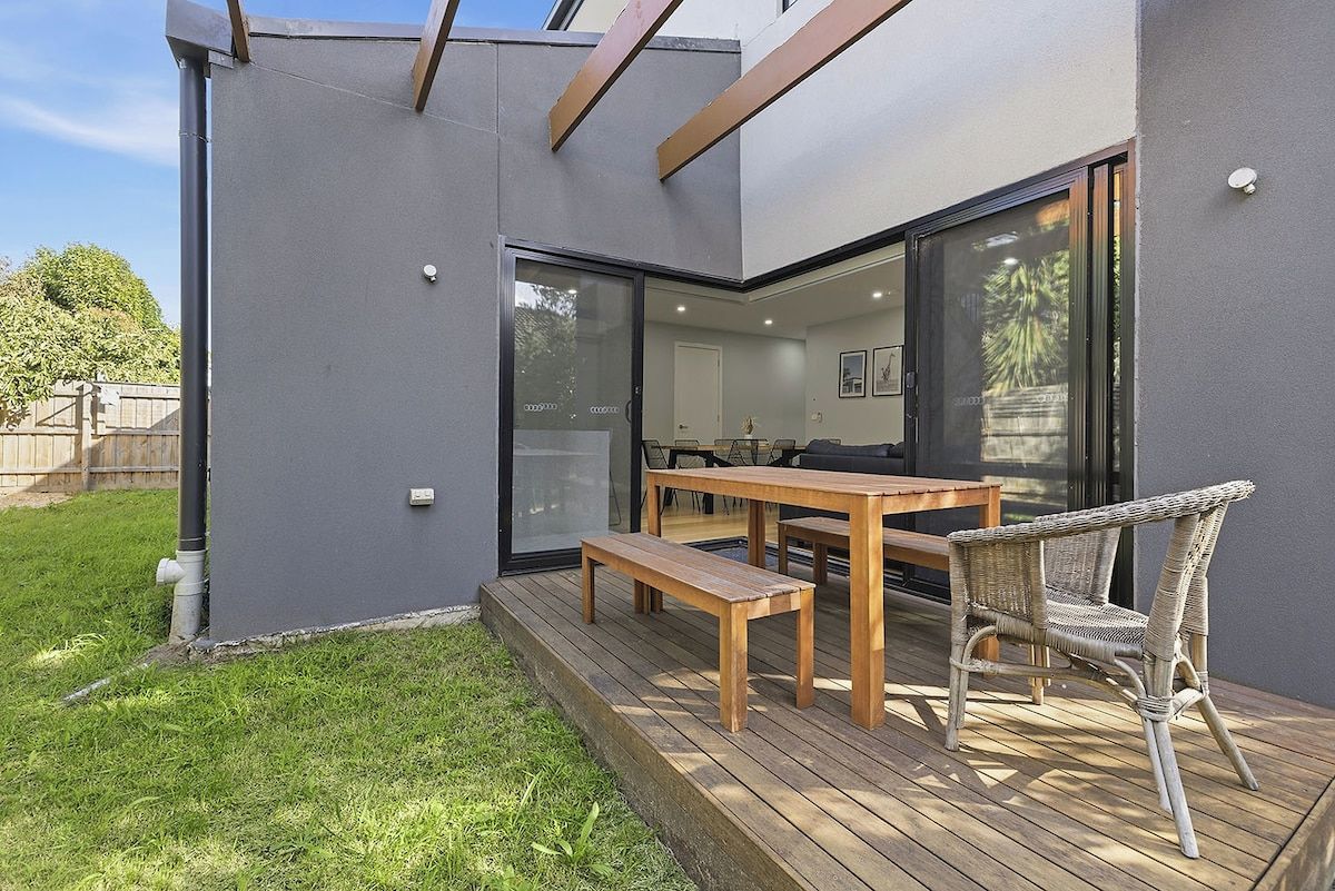 A modern outdoor patio with a wooden table and chairs, a glass door leading to an interior space, and a grassy area.
