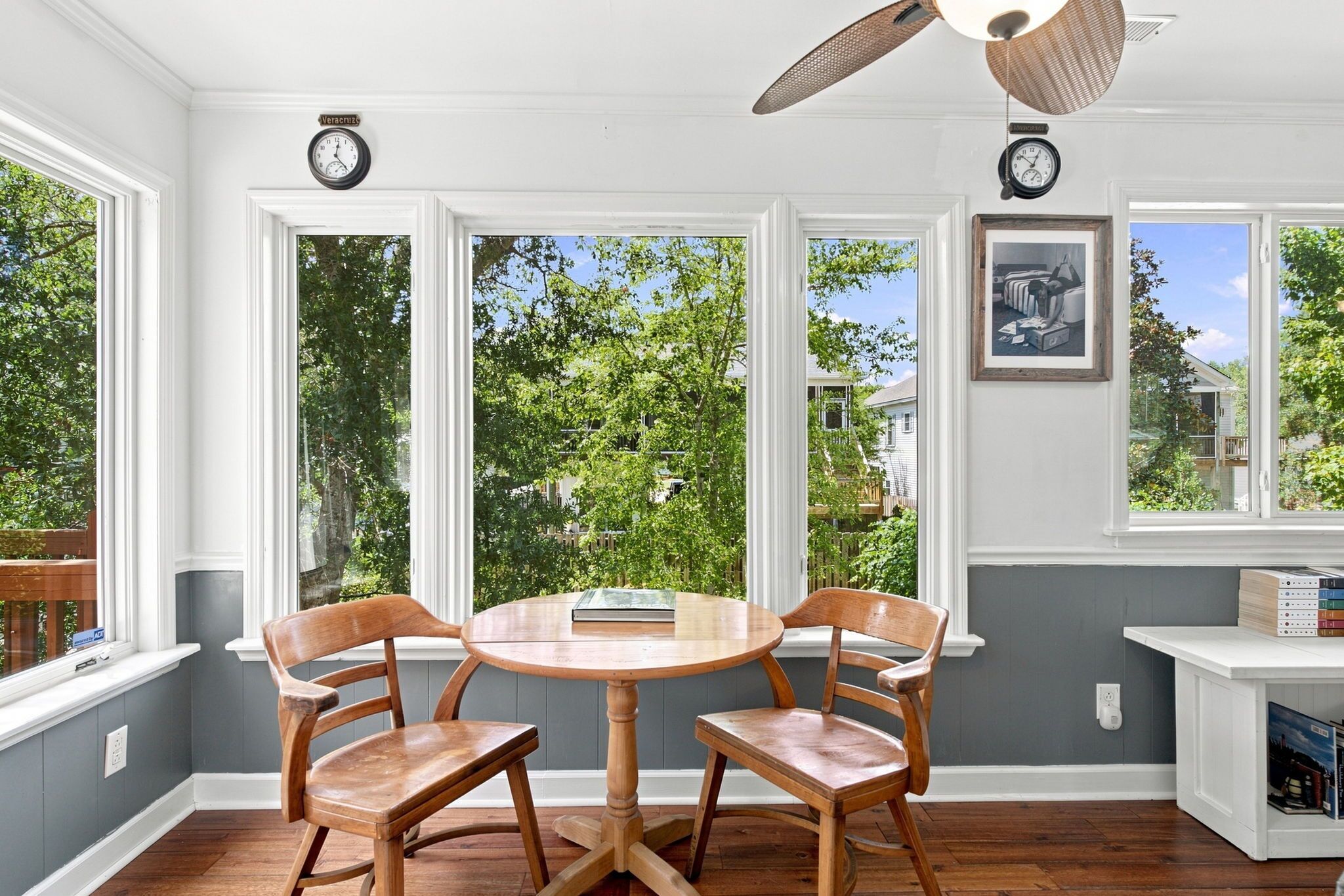 A dining area with a round table and four chairs, large windows with a view of greenery, and a ceiling fan.