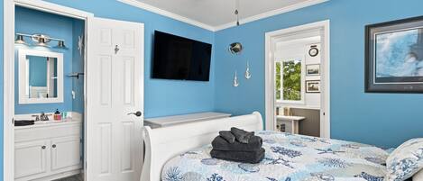 A bedroom with a bed, a television, a ceiling fan, and a door leading to a bathroom.