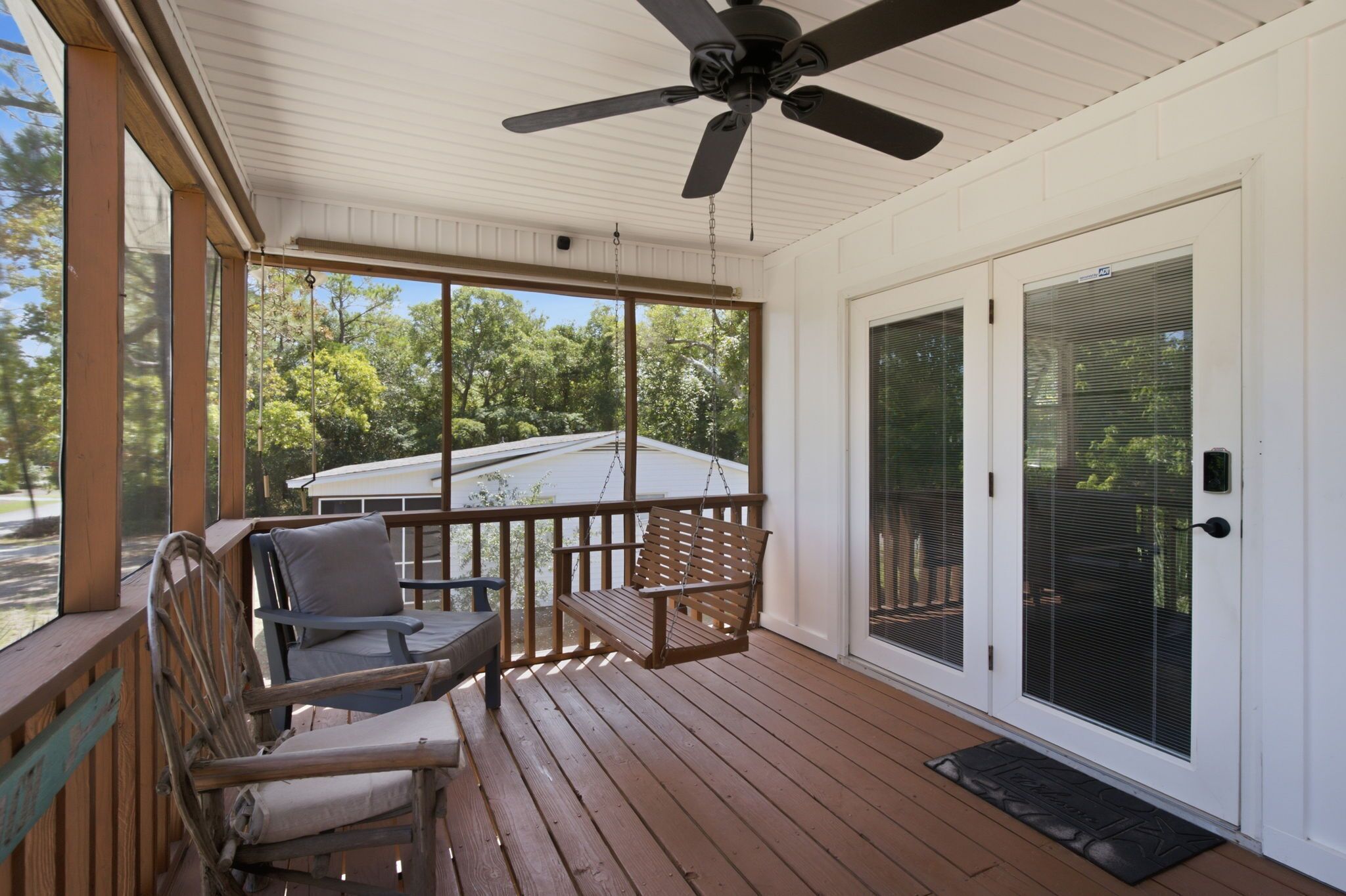 A covered porch with a ceiling fan, two chairs, a swing, and a sliding glass door.