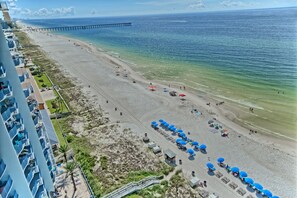 Ein Strand mit einer Mole, einem Hotel mit Balkonen und Strandliegen.