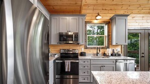 A modern kitchen with stainless steel appliances, wooden ceiling, and a window providing a view of greenery. - Nature’s Ridge | Modern Cabin + Stunning Views (Sevierville)
