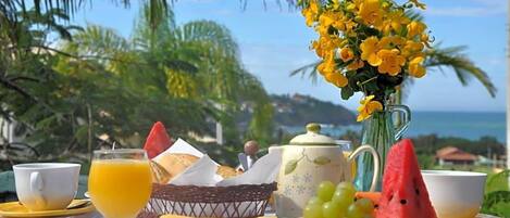 A breakfast table with a variety of food items, a vase of yellow flowers, and a view of the ocean.