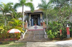 A house with a staircase leading to a door, surrounded by lush greenery and a palm tree. - Ferradura Parque Pousada (Búzios)