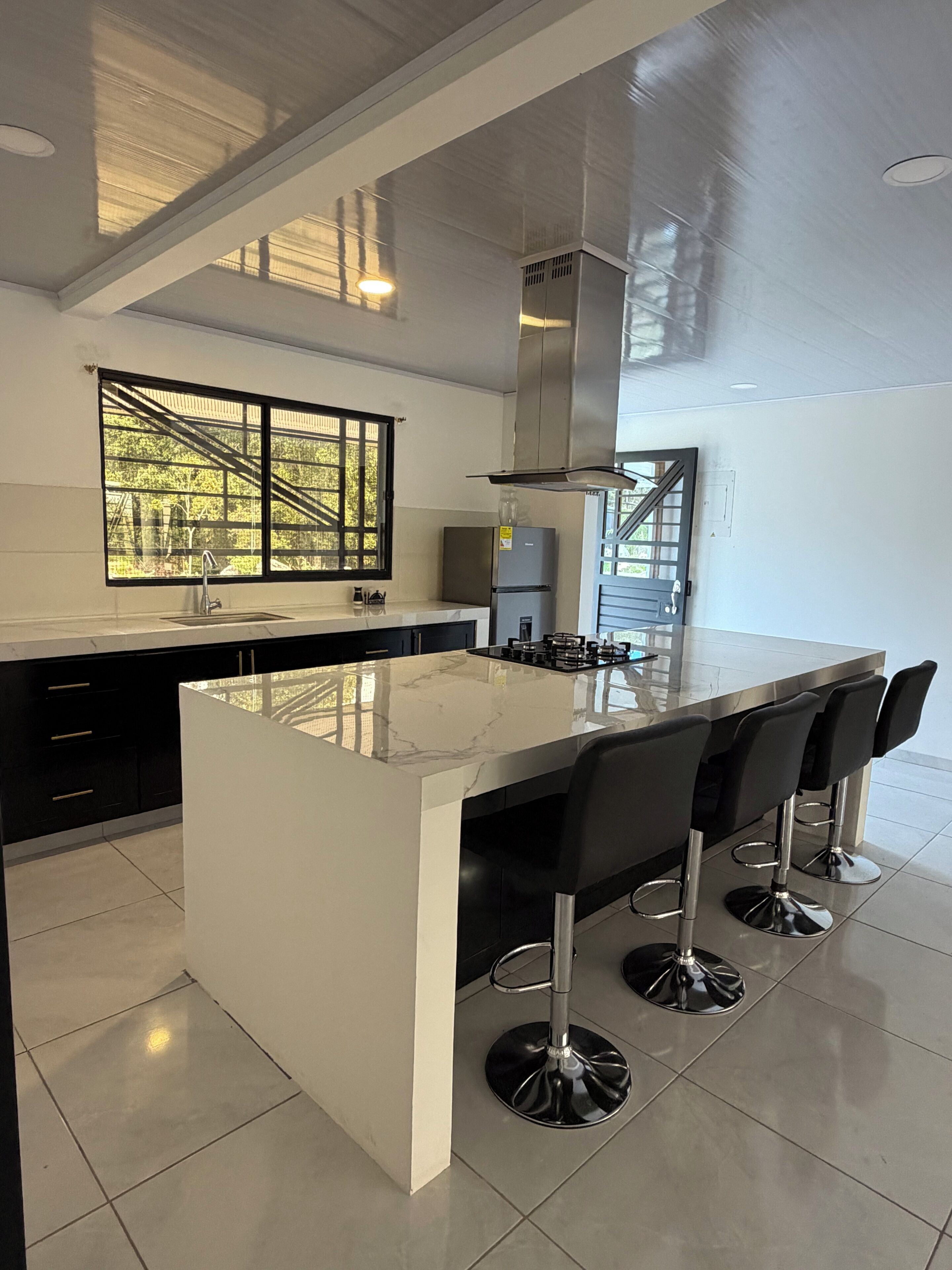 A modern kitchen with a large island, bar stools, a stainless steel refrigerator, and a window with a view of greenery.