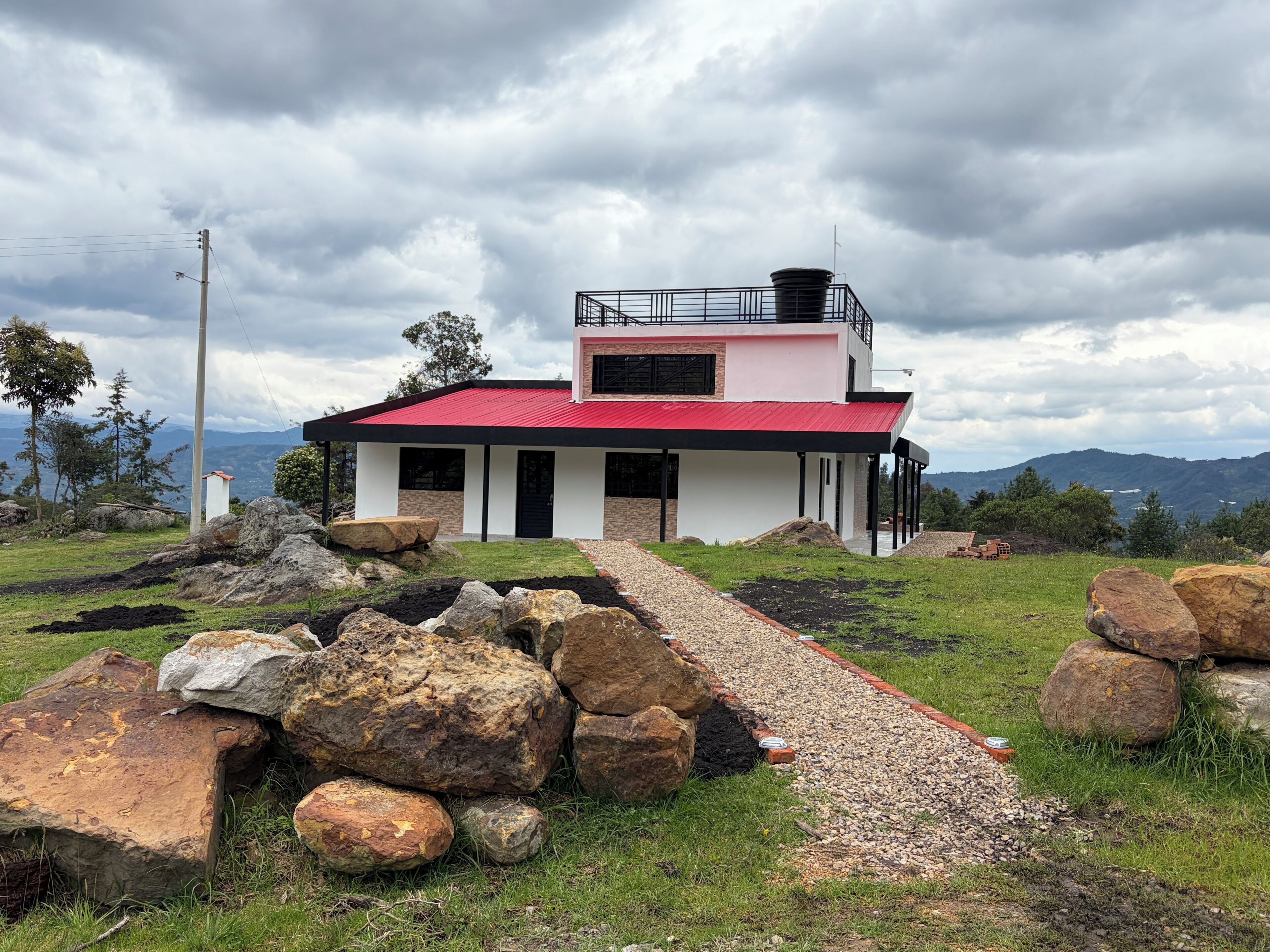 Une maison moderne avec un toit rouge, entourée de gros rochers et bordée d’un chemin de gravier.