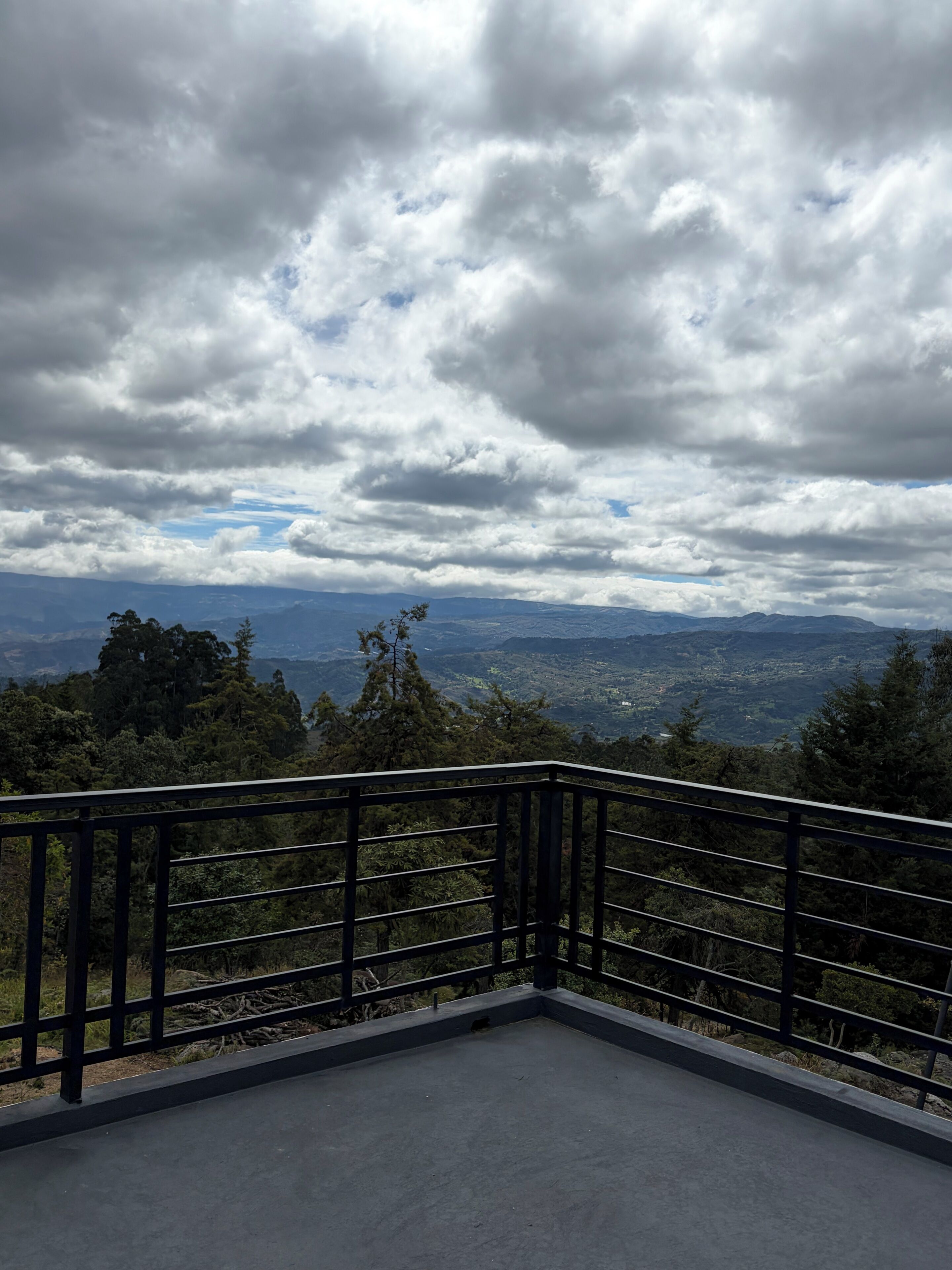 Un balcón con baranda de metal que da a un paisaje montañoso con vegetación densa y cielo nublado.