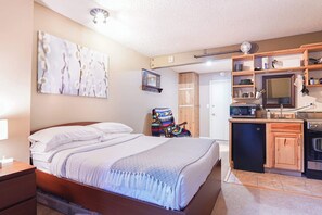 Une chambre à coucher comprenant un lit, une table de chevet, un petit bureau, une chaise, une armoire, et un coin cuisine équipé d’un four à micro-ondes, d’un évier et d’un réfrigérateur.