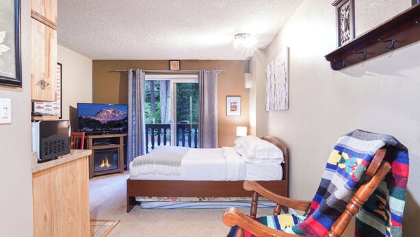 A bedroom with a bed, a television, a fireplace, and a balcony with a view of trees. - Fireside Suite, Simple Basecamp at Snowline Lodge (Deming)
