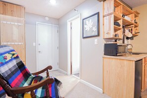 A compact kitchen with wooden cabinets, a microwave, and a chair with a colorful blanket.