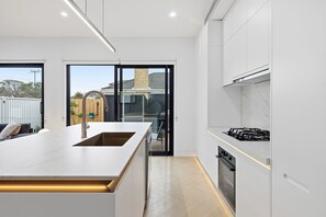 A modern kitchen with a large island, built-in appliances, and a sliding glass door leading to a backyard. - Domi Rentals - The Cheltenham Southland House (Cheltenham)