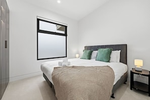 A modern bedroom with a large bed, a nightstand, and a wall-mounted mirror. - Domi Rentals - The Cheltenham Southland House (Cheltenham)
