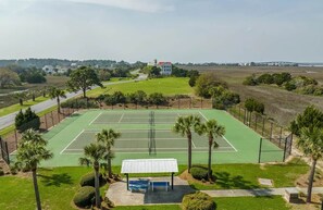 Tenniskenttä, jossa on katettu leikkimökki, ja kentän ympärillä on palmuja sekä näkymä kauempana sijaitseviin rakennuksiin.