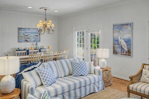 A living room with a striped sofa, a dining area with a white table and chairs, a chandelier, and artwork on the walls.