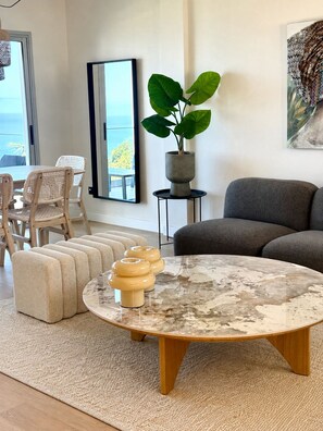 Ein modernes Wohnzimmer mit einem runden Couchtisch, gepolsterten Hocker, einer Zimmerpflanze und Blick ins Freie.