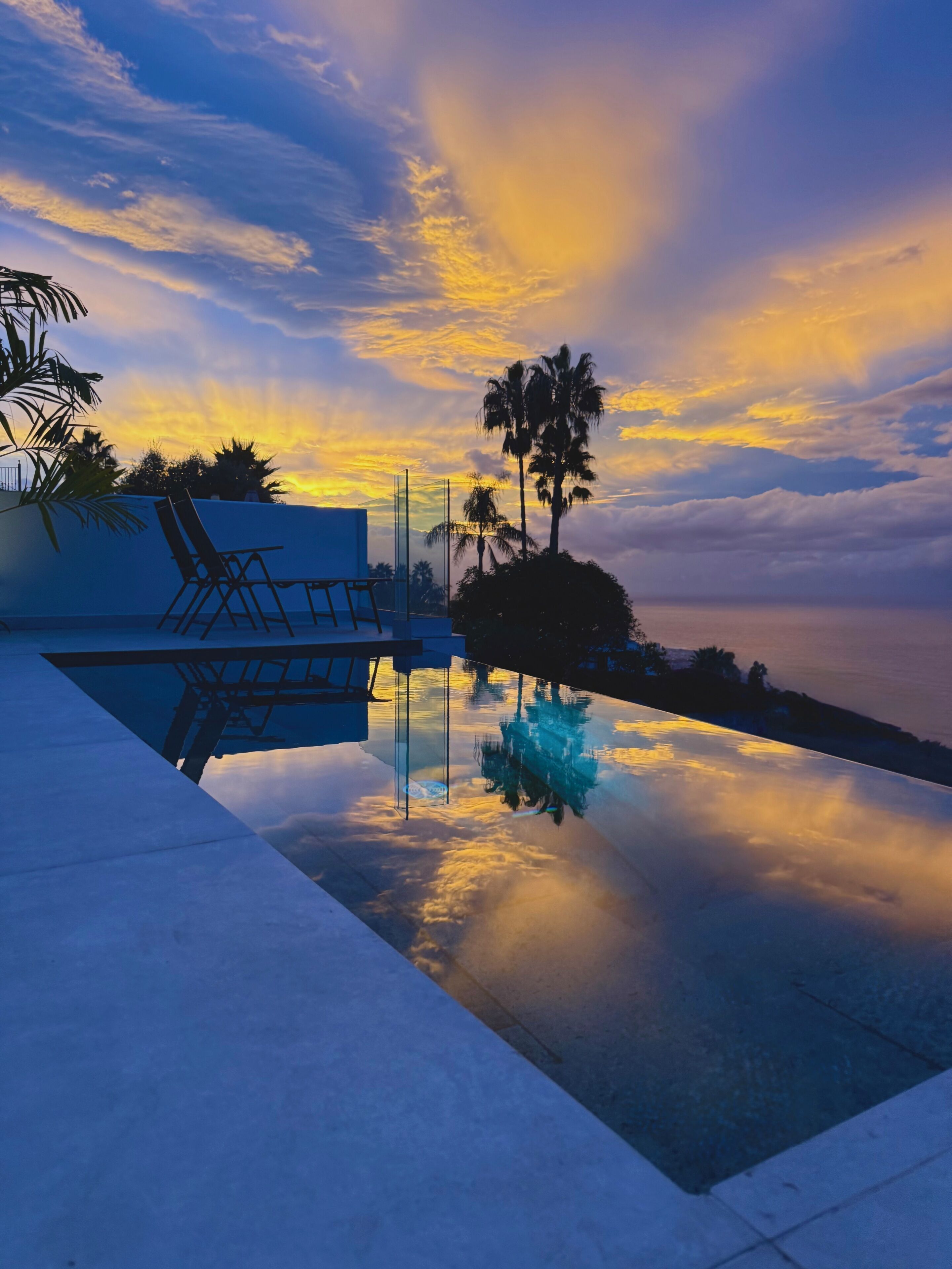 Ein Pool, der Himmel, Palmen und ein Stuhl sich darin spiegeln.