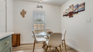 Een eetgedeelte met een glazen tafel, witte stoelen, wanddecoratie met een cactus, een raam met zonwering en een plafondlamp.