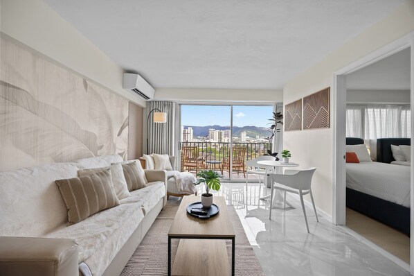 A modern hotel room with a sofa, a coffee table with a plant, a dining area with a table and chairs, and a view of the cityscape through large windows. - Water View, Corner 2 bedroom Condo in Waikiki, Free Parking (Honolulu)
