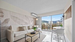 A modern living room with a sofa, coffee table, and a balcony with a view of buildings and mountains. - Free Parking & Beach Gear at 2BR Iconic Whale Building Condo (Honolulu)