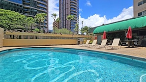 Ein Swimmingpool mit Liegestühlen und Sonnenschirmen, umgeben von Gebäuden und Palmen.