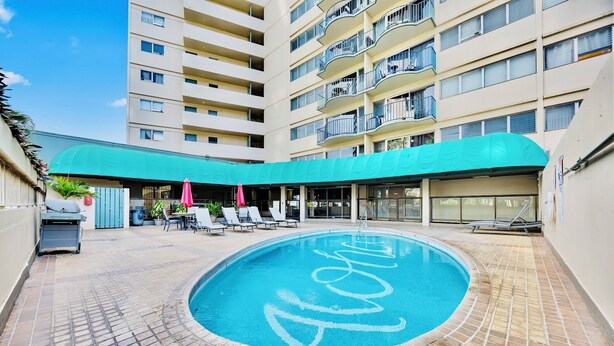 A swimming pool with a tiled deck, surrounded by lounge chairs and umbrellas, with a multi-story building in the background. - Free Parking & Beach Gear at 2BR Iconic Whale Building Condo (Honolulu)