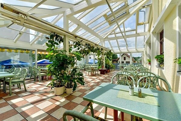 A covered patio area with tables and chairs, potted plants, and a glass ceiling structure. - Nagerl Landhotel (Marzling)